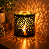 A black metal scented candle holder with a tree of life cutout design, lit with a warm glowing candle inside. The holder sits on a wooden table, casting a detailed leaf pattern shadow onto a wall behind it.