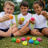 Three children sitting on grass holding colorful smiley face balls with more balls in front of them.